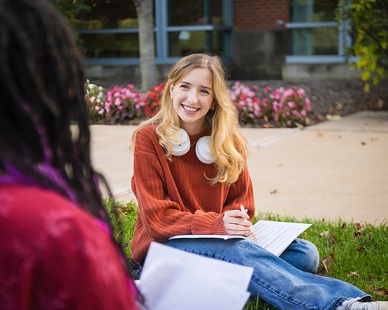 A student sitting on University lawn with headphones around her neck, talking with someone in the foreground.