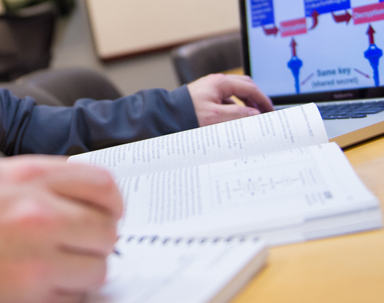 Student's hands turning pages of an open textbook with laptop nearby